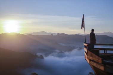 Huzurlu adam, ruhani düşüncelere dalmış keşiş güzel bir güneşin doğuşunu izliyor. Kalın sisle kaplı engin dağ manzarasına bakan ahşap bir manzaranın üzerinde duruyor.