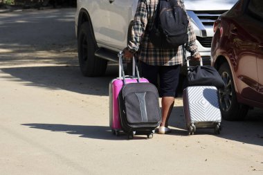 Asfaltsız bir yolda bavul ve pembe bavul taşıyan tanınmamış bir gezgin. Sırt çantalı ve çantalı kişiler arabanın yanında dururlar, tatile çıkarlar ya da yolculuğa çıkarlar.