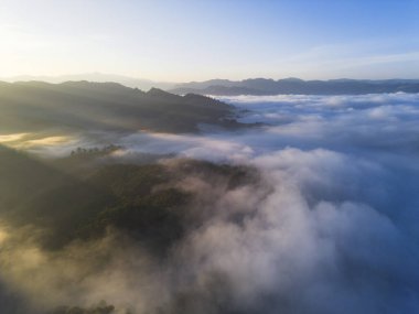 Güneşin doğuşuyla birlikte bulutlar üzerinde sakin bir dağ manzarası. Altın güneş ışığı siste parlar, yolculuk için huzurlu ve görkemli bir arka plan yaratır.