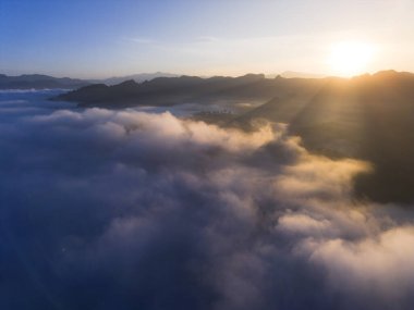 Dağ sırasının üzerinde güzel bir gün doğumu manzarası. Sis denizi aşağıdaki vadiyi kaplıyor, altın güneş ışığı ve berrak mavi gökyüzü huzurlu bir manzara yaratıyor.