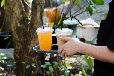 Server, serinletici soğuk içecekler içeren siyah tepsi tutuyor. Soğuk kahve, meyve suyu ve açık hava bahçesindeki bir kafede taze hindistan cevizi gibi.