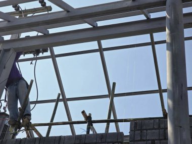 İnşaat işçisi çelik çatı iskeleti üzerinde çalışıyor. Çalışkan adam açık mavi gökyüzü altında el işçiliği inşa yapısını gösteriyor