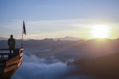 Tahta perspektifteki yalnız adam güneşin doğuşunda sakin dağ manzarasının tadını çıkarıyor. Düşünceli gezgin, sis ve bulut denizinin üzerindeki güzel doğa manzarasına bakıyor.