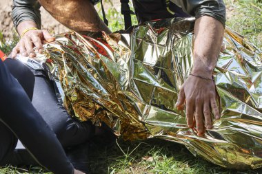 Kurtarma ekibi, acil durum battaniyesiyle kaplanmış yaralı kişiye dokunuyor. Kriz anında yardım ve destek gösteriyor.