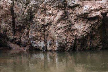 Durgun nehir suyunun üzerinde beliren engebeli kahverengi uçurum dokusu sakin doğal kanyon sahnesinde taş yüzeyini yansıtıyor. Huzurlu ruh hali uyandırıyor.