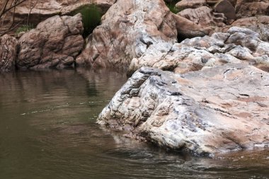 Durgun nehir suyundan yükselen geniş desenli kaya oluşumuna sahip huzurlu doğal manzara. Detaylı taş yüzeyli vahşi ortamda sakin bir atmosfer yaratıyor.