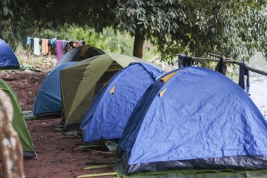 Mavi ve yeşil kamp çadırları kamp alanındaki kırmızı toprak zeminde sıralanmıştı. Arka planda giysiler kuruyordu. Dışarıdaki gündüz macerası sırasında sakin doğa atmosferini gösteriyordu.