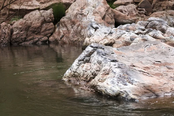 Durgun nehir suyundan yükselen geniş desenli kaya oluşumuna sahip huzurlu doğal manzara. Detaylı taş yüzeyli vahşi ortamda sakin bir atmosfer yaratıyor.