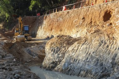 Kazıcı, yüksek toprak duvarlı çamurlu akarsuyun yanında toprak kazıyor. Ağır makineler ve toprak işçiliğini içeren büyük inşaat projesinde kararlılık gösteriyor.