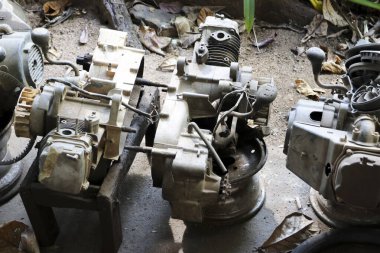 Parçalanmış motor parçaları metal dişliler ve kovanlarla kirli tamir atölyesinin zemininde yatıyor. Endüstriyel grilik ve terk edilmiş bakım hissi uyandırıyor.