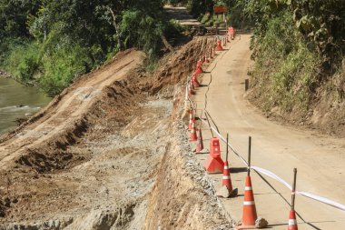 Turuncu güvenlik konisi işaretleriyle hasar gören kırsal yol, dik toprak kayması ve nehrin kenarında tehlikeli kenar çizgileriyle onarılıyor. Bu da seyahat taşıma inşaat alanının güvenliği için uyarıda bulunuyor.