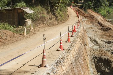 Toprak yolda inşaat çalışması. Uçurumun kenarında güvenlik konisi var. Güneşli bir günde bitmemiş kırsal bir yol var.
