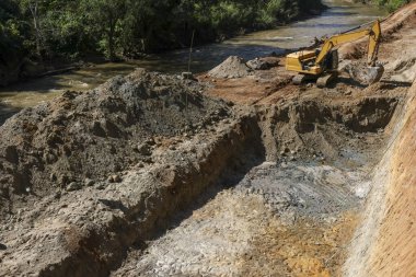 Aktif sarı kepçe kazıcı nehir kıyısının yanında çamurlu ve kumlu inşaat alanı için büyük bir çukur kazıyor.