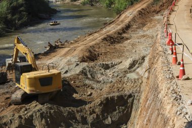 Ağır sarı kazıcı makineleriyle kazı alanı nehir kıyısında toprak kazıyor ve güvenlik konileri tehlikeli bölgeyi koruyor endüstriyel gelişim ve mühendislik ilerlemelerini gösteriyor.