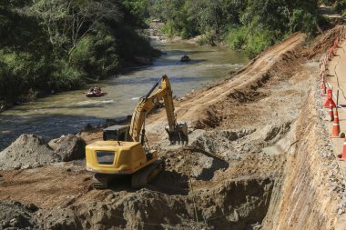 Nehir kıyısı şantiyesinde toprak kazan ağır sarı kazıcı güçlü mekanik kollarla akan su ve yeşil orman arka planının yanı sıra altyapıyı geliştiriyor.