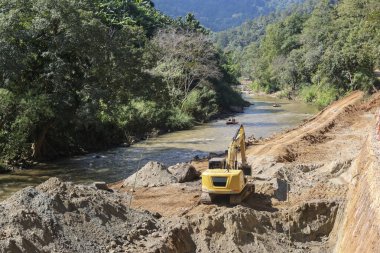 Ağır sarı kazıcı toprak kazıyor sakin nehir kıyısı inşaatı umut verici bir ilerleme ve gelişim atmosferi yaratıyor ağaçlar ve dağ altyapısı ile uzak doğa ormanı manzarası