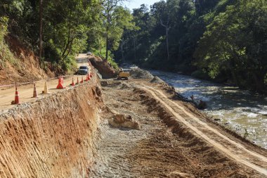 Orman manzarasının ortasında akan nehir kenarındaki bitmemiş kırsal yolun yer aldığı inşaat alanı altyapı gelişimini ve doğal çevrenin dönüşümünü gösteriyor