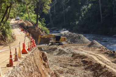 Sarı kazıcının nehir kıyısında toprak kazdığı, turuncu trafik konilerinin toprak yola dizildiği inşaat sahası, altyapı gelişiminin yoğun ilerleyişini, kırsal çevre koşullarını ifade ediyor.