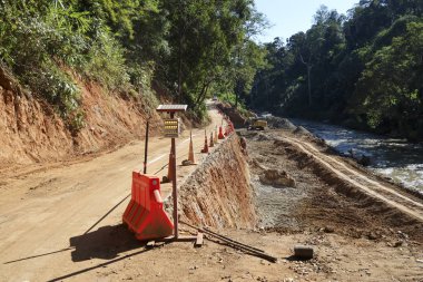 Kirli bariyerlerle dolu yol inşaatı ve nehir kıyısında kırsal orman manzarasında tamamlanmamış patika karmaşık gelişim ve ilerlemeyi gösteriyor.