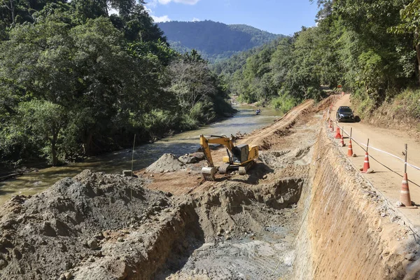 Nehir kıyısında toprak kazan kazı yapan bir inşaat arazisi. Dağ ormanlarının arka planında engebeli arazi ve doğada sakin bir atmosfer görünüyor.