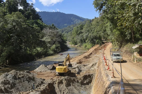 Nehir şantiyesi yakınlarında toprak kazan ağır sarı kazıcı, yeşil dağ arka planında endüstriyel ilerlemeyi ve uzak doğal ortamda yol yapımı için muazzam toprak çalışmalarını gösteriyor.