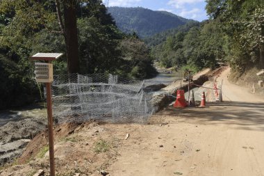 Güneşli bir günde mavi gökyüzünün altında yeşil orman arkaplanı yakınlarındaki metal tel örgüden yapılmış tozlu yol koruma çitlerinin yanında akan nehir akıntısı ile huzurlu kırsal alan.