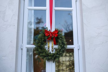 Geleneksel yeşil Noel çelengi beyaz pencere çerçevesinde asılı. Mutlu bayram sezonunu kutluyor. Kış kutlaması için neşe ve karşılama ruhu katıyor.