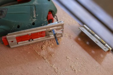 Yakın plan, odaklanmış marangozluk ev yenileme projesi sırasında gümüş metal menteşenin yanındaki talaş tozuyla kaplı ahşap yüzey üzerinde duran yeşil elektrikli yapbozu gösteriyor.