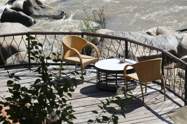 Boş balkon, hasır sandalyeler ve çalkantılı nehir akıntısı ile dolu bir masa. Huzurlu, sakin bir atmosferi olan kaya taşları arasında akıyor.
