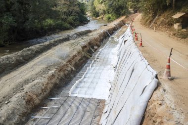 Yol kenarı takviyesi, nehir kıyısına yakın beyaz jeotextile kumaş ve turuncu koni ile uzak doğa alanında ilerleme ve gelişme gösteriyor.