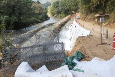 İnşaat alanı Gabion duvar yapısını gösteriyor. Kırsal doğa bölgesindeki hasarlı altyapıyı koruyan jeotektil kumaş ve güvenlik konileri ile yol kenarındaki nehir kıyısını güçlendiriyor.