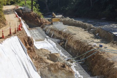 Nehir kıyısı boyunca inşaat sahasında kazılmış toprak ve beyaz kum torbasıyla yapılan koruma çalışmaları endüstriyel ilerlemeyi ve doğanın gelişimini gösteren bir duvar oluşturur.