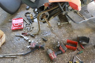 Kırık aracın yüksek açılı çekimi ıslak beton zeminde kirli parçalarla mekanik aletleri ve kaotik garaj atmosferinde paslı dörtlü bisikleti gösteriyor.