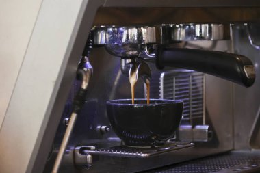 Profesyonel espresso makinesi koyu bardağa taze kahve demliyor. Klasik sabah rutinini ve kafenin içindeki metal portafilterden dökülen aromatik sıvı akışla barista sürecini gösteriyor.