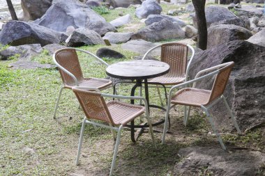 Boş sandalyeleri ve yuvarlak masaları olan açık hava mobilyaları bahçedeki yeşil çimlerin üzerinde, büyük doğal kaya ve ağaçlarla çevrili.