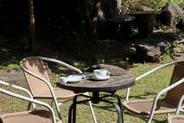 Sakin bir sabah tatili için güneşli doğa parkı için mükemmel bir kahve fincanı tabağı ile küçük ahşap masanın yanında dinlendirici metal koltuklar içeren açık hava kafesi.