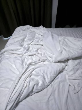 Yatak odasındaki tembel sabah atmosferini yansıtan buruşuk yorgan ve dağınık çarşaflı beyaz yatağı yakın çekimde göster.