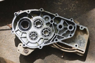 Kirli metal motor parçası, bilye konut mekanizması ve motor bakımından kalma yağlı krankcase kapağı sert beton arka planda endüstriyel aşınma ve yırtık dokusunu yansıtıyor.