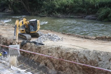 Sarı kazıcı makinası ağır ekipman nehir kıyısında toprak ve taş kazıyor. İnşaat alanı için endüstriyel dayanıklılık ve gelişim bulutlu bir günde hissediliyor.