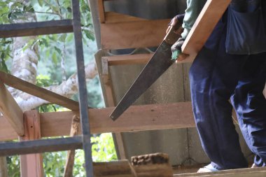 Büyük bir el testeresi tutan usta bir inşaat işçisi, ahşap kiriş iskelet yapısı ile marangozluk inşaatına odaklanmış enerjik tavrıyla dışarıda çalışkan bir işçilik sergilemektedir.