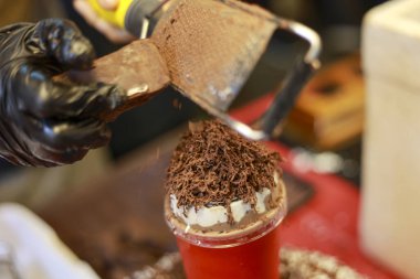 Profesyonel şef, siyah eldivenli, çikolatayı rendeleyen, kremalı, metal rende kakao bardağıyla lezzetli tatlı içeceği yapan.