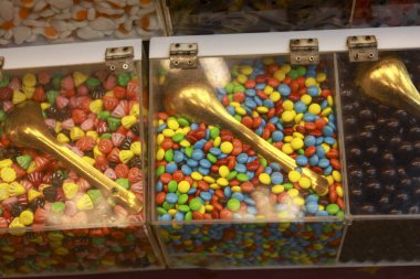 Çikolata kaplı renkli şekerlemeler, altın kepçesiyle aç müşteriyi lezzetli atıştırmalıklar için perakende satış tezgahındaki akrilik konteynır vitrininden lezzetli bir ikram seçmeye davet ediyor.
