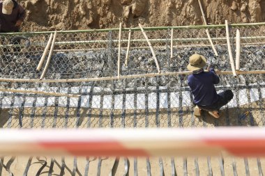 Tel örgülü Gabion duvar yapısına çömelmiş inşaat işçisi. Açık havada ciddi bir ifade takınıyor. Parlak güneş ışığı altında toprak zemini projesinde çalışıyor.