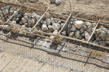 Kaba taş yığınıyla dolu bir temel siperinin yer aldığı inşaat sahası. Parlak güneş ışığı altında dayanıklı çelik inşaat iskeletiyle güçlendirilmiş. Beton dökme hazırlıklarını gösteriyor.