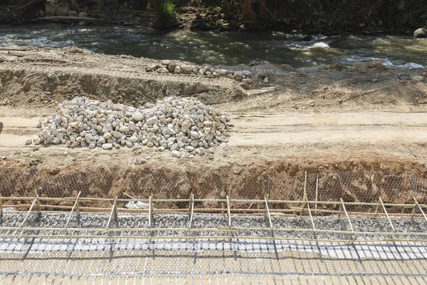 Parlak güneşin altında taş yığınları ve kuru toprak yığınlarıyla nehir kıyısına yakın çelik inşaat demiri ızgarası ile güçlendirilmiş beton temele sahip inşaat alanı