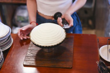 Odaklanmış kadın pasta şefi beyaz yuvarlak pastayı dikkatlice dekore ediyor. Mutfak dükkanının içindeki ahşap masaya meşale koyuyor. Ciddi bir bağlılık tutkusu var.