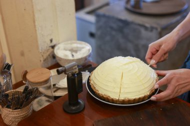 Elleri kaymaklı limonlu turtayı ahşap masada kesiyor. Fırın aleti ile dikkatli bir hazırlık ve tatlı bir his veriyor.