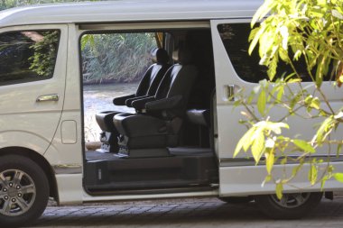 Açık kapılı beyaz yolcu minibüsü, turistleri bekleyen yeşil ağaç yapraklarının altına park edilmiş içi boş koltukları gösteriyor.
