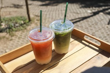 Buzlu çay ve temiz plastik bardaklarda servis edilen soğuk kibrit latte, tahta tepside pipetlerle güneşli bir günde dışarıda serin ve rahatlatıcı bir yaz havası sunuyor.