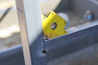 Sarı manyetik kaynak aleti, iki parça çelik boruyu dik açı köşesinde tutuyor. İnşaat alanının arka planında odaklanmış ve hassas üretim havasına sahip kaynaklara hazır.